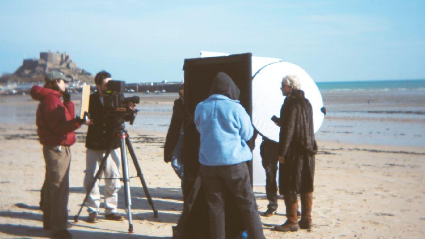 Film crew and cast stand on a beach while filming. A camera man is operating a camera which is pointing at cast members. It's a sunny day.