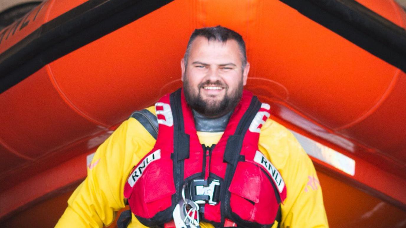A portrait photo of Lee Stothard smiling at the camera. He is wearing a yellow jacket and a red harness with various equipment. He has short brown hair and beard. His eyes are blue. 