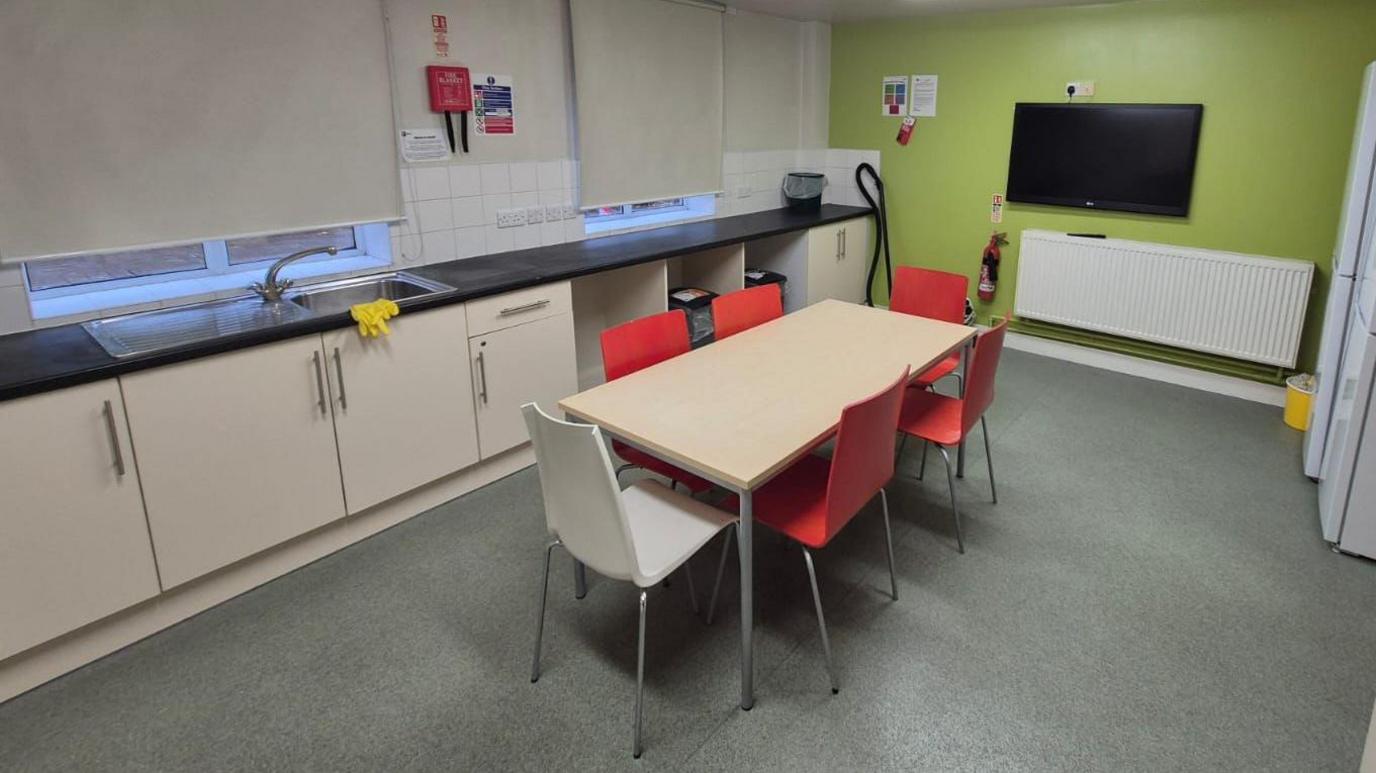 A kitchen with a table, four red chairs, a white chair and a green wall with a television on it. There is a white radiator and kitchen cupboards. There are fridges on the right. The floor is a greenish-grey. Blinds are pulled down at the two windows.