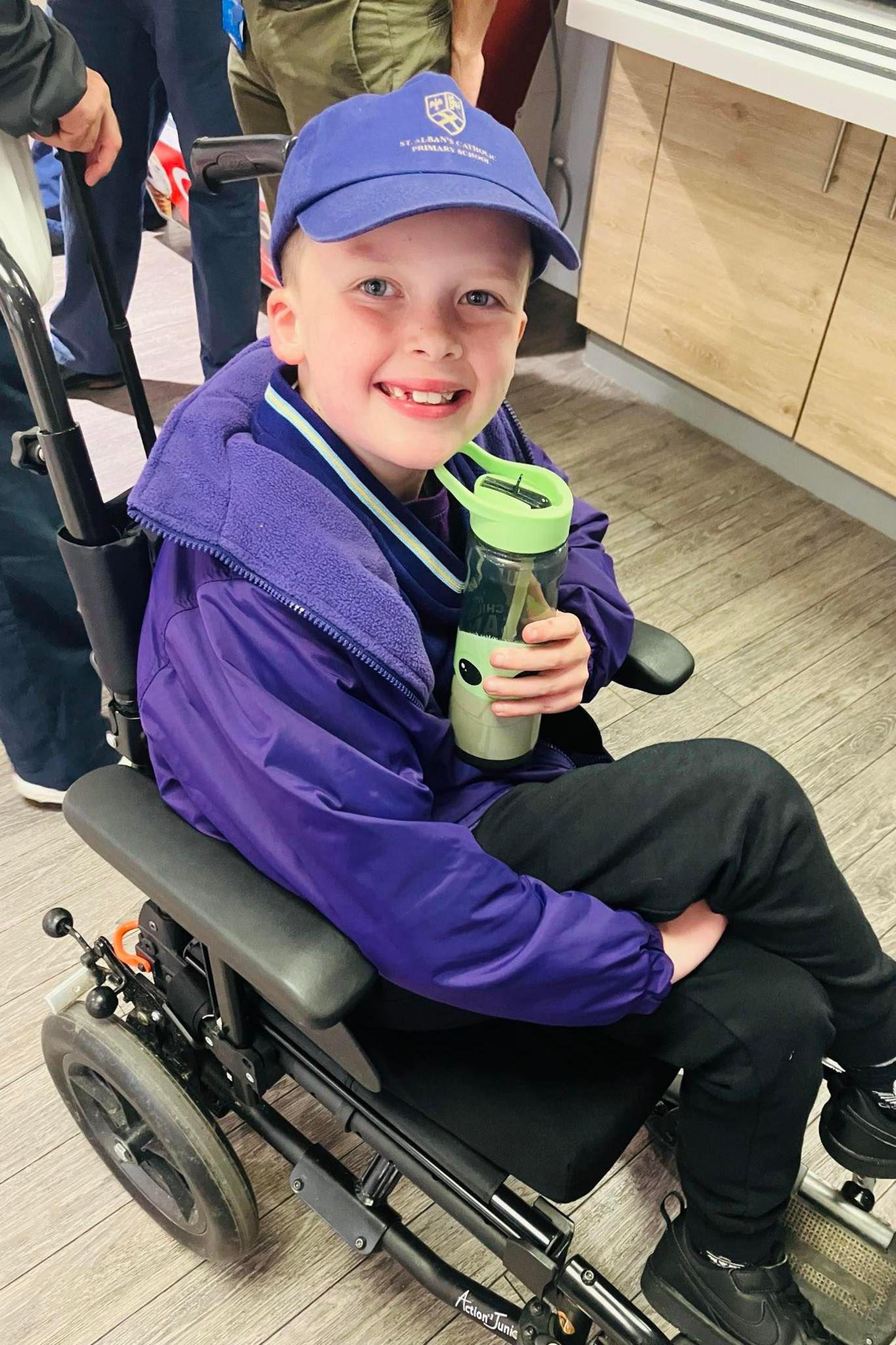 Vinnie, wearing a purple jacket and cap, sitting in a wheelchair, holding a water bottle and smiling up at the camera.