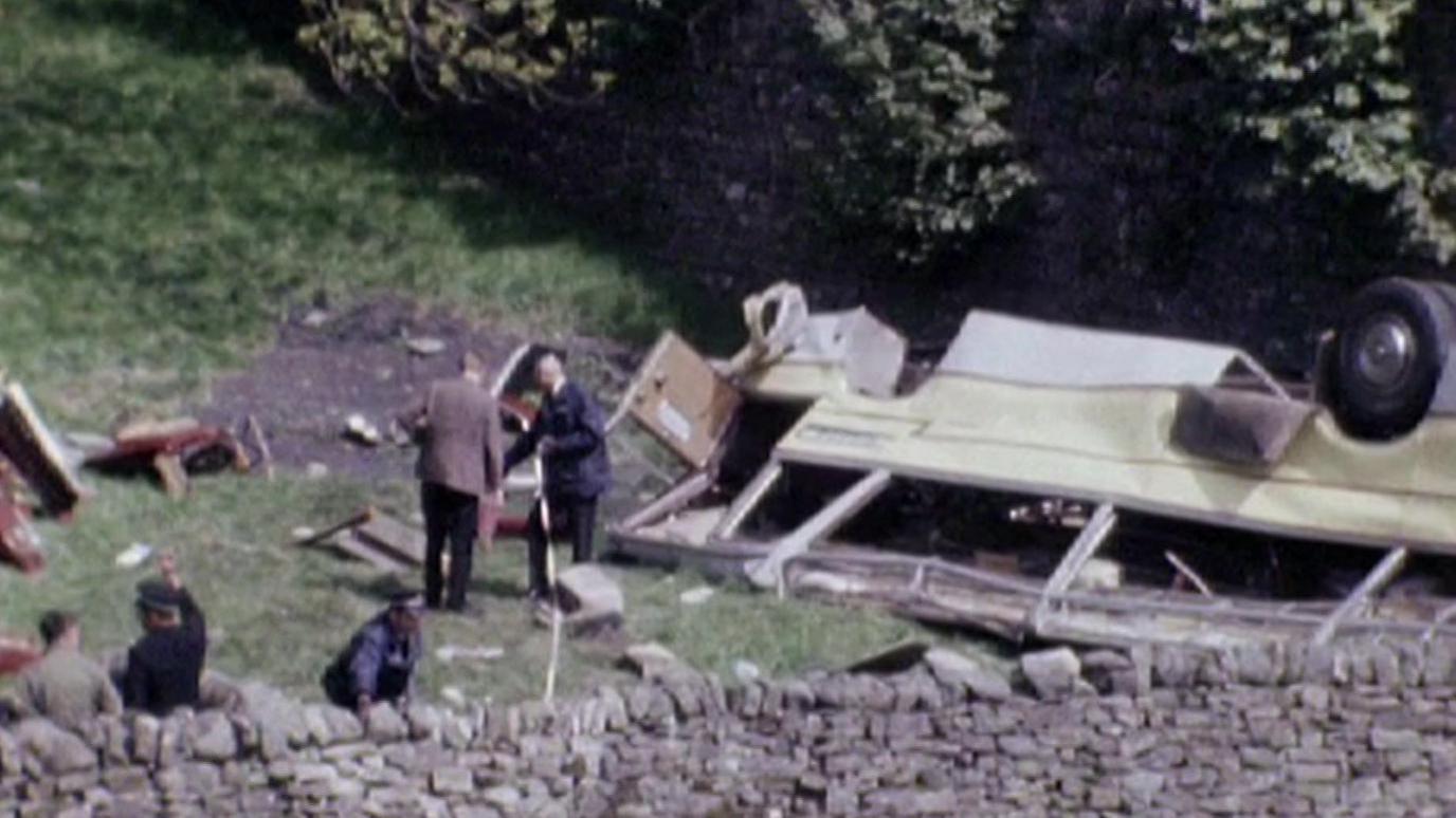 A group of men inspect the wreckage of a coach crash. They stand against a dry stone wall