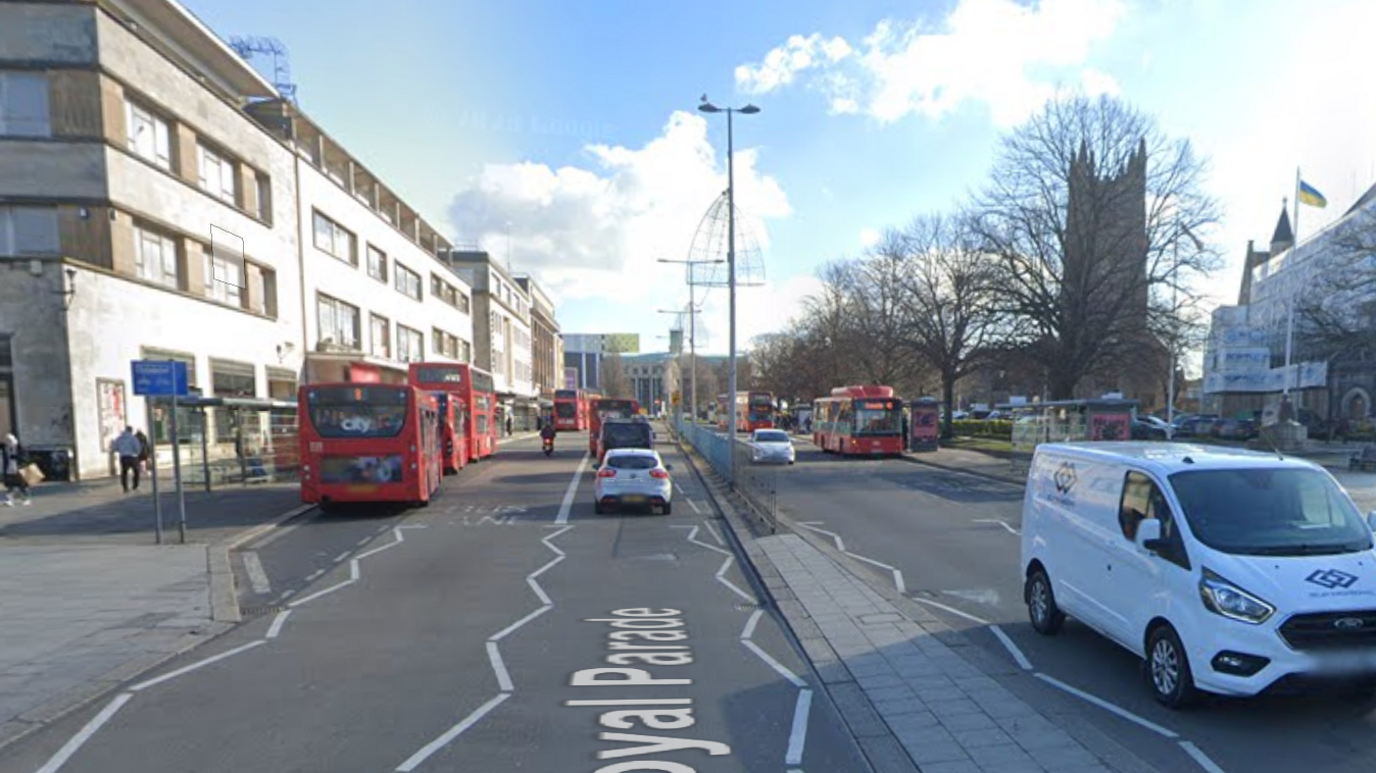 A google street view image of Royal Parade Plymouth. On the left had side of the road there are shopping buildings. On the road there are two buses and one double-decker bus stopped at a bus stop. There are cars and buses driving on the road. On the right there are two buses stopped at bus stops, front towards the camera. There is a church. 