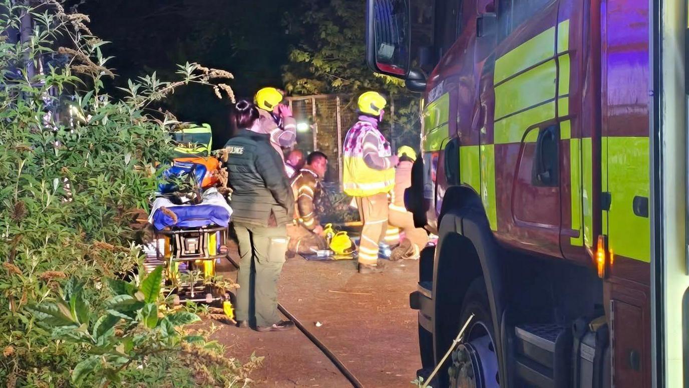 A paramedic with an ambulance bed stood alongside five firefighters by the front of a fire engine. Three of the firefighters are knelt on the ground. The paramedic looks on.