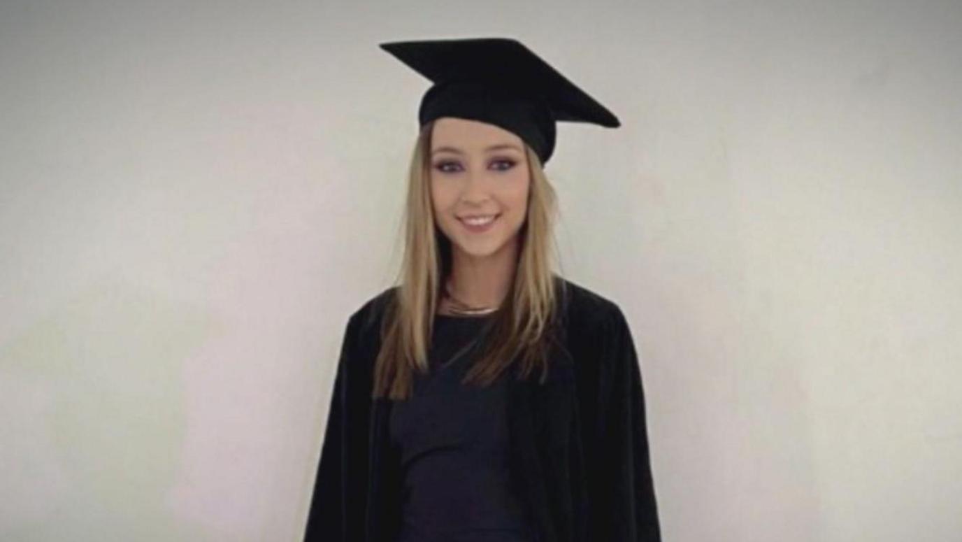 Jessica Gallagher smiling at the camera on her graduation day.  She is wearing a black mortarboard and a black robe over a black or dark navy dress.  She has long, straight blonde hair and is standing in front of a plain, pale background. 