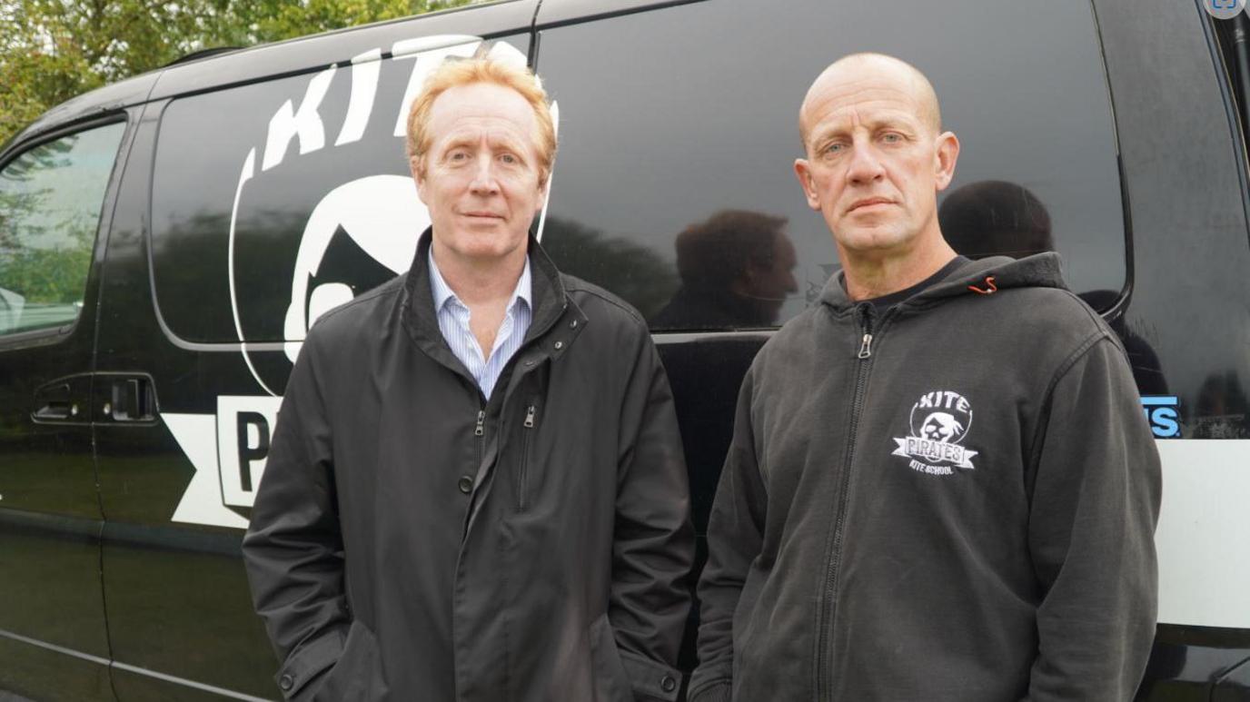 Stefano Biagini and Graeme Rolbieckie are both dressed in dark grey jackets. They are standing in front of a dark grey van with a logo in white, which is partially covered, but the word Kite is readable