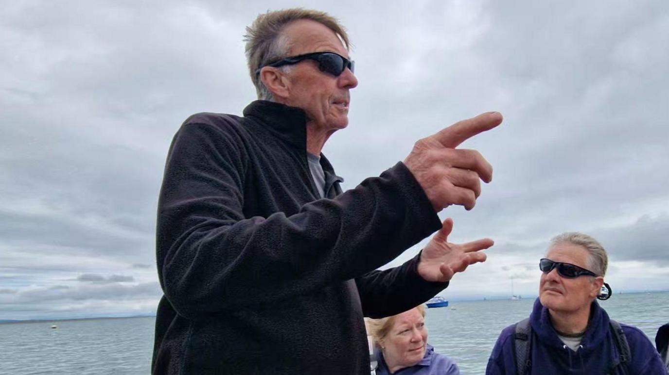 Steve Wilcock standing on his boat looking out to the sea and speaking to passengers who are seated on the vessel. He is portrayed side-on and he is pointing at something with one hand. He has short grey hair and he is wearing a lack fleece and dark sunglasses.