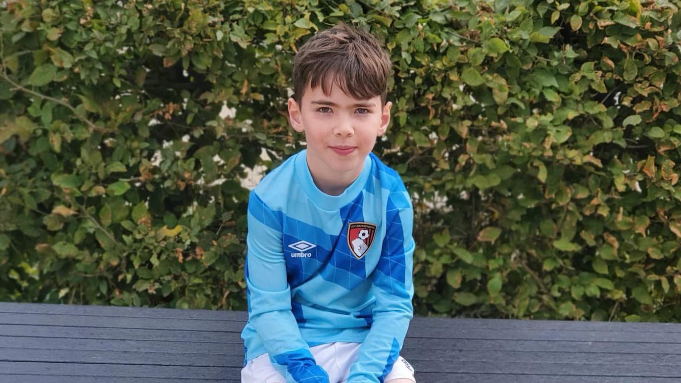 Reuben is sitting on a bench outdoors, surrounded by lush green foliage. He is wearing a blue Umbro sports jersey with geometric patterns and an AFC Bournemouth football club emblem. He has short brown hair and is looking directly at the camera.