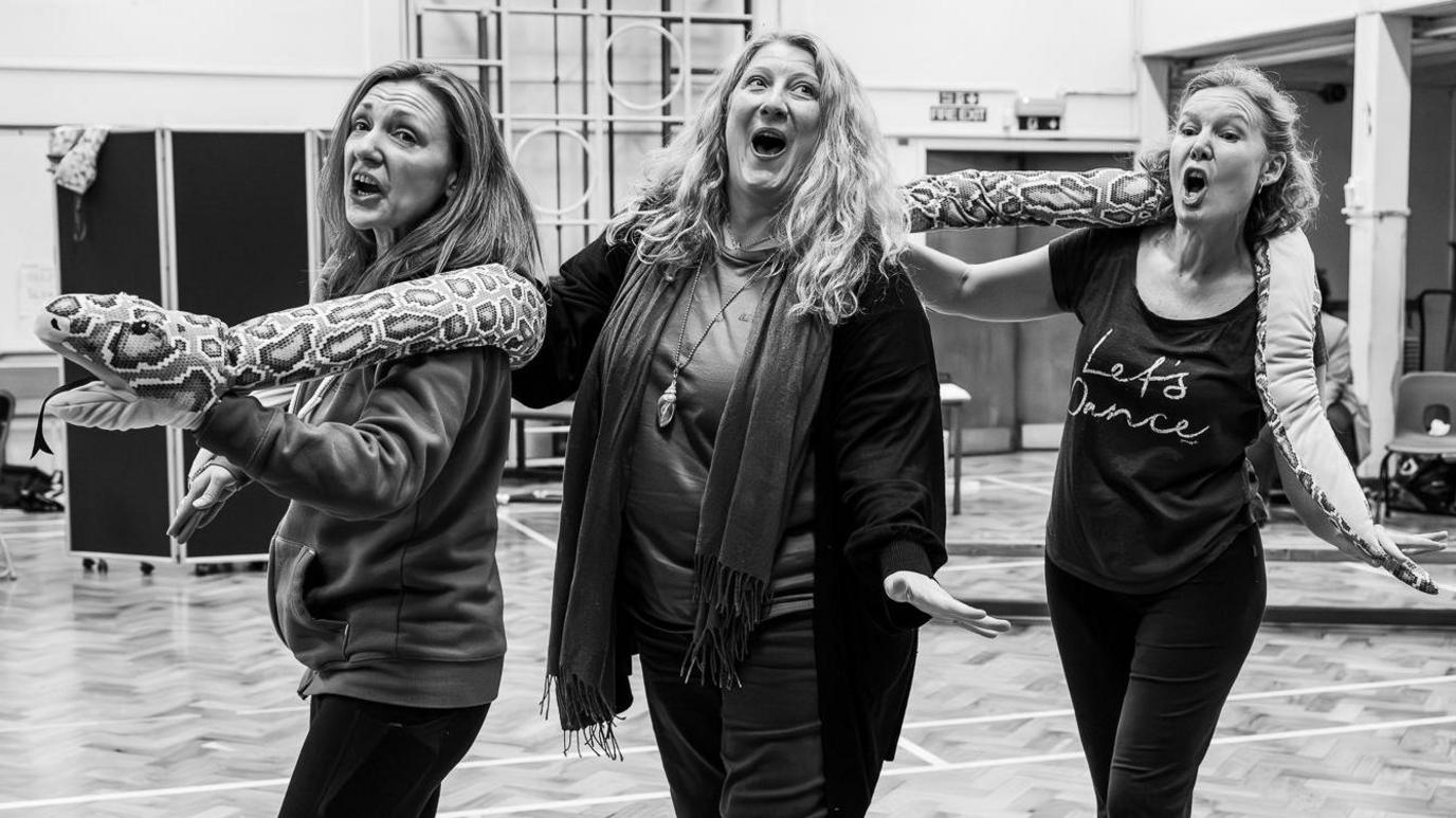 Three women taking part in a rehearsal. Their mouths are open and their arms are out and they are holding a toy snake. The image is black and white and the rehearsal is taking place in a wall with sport equipment behind. 