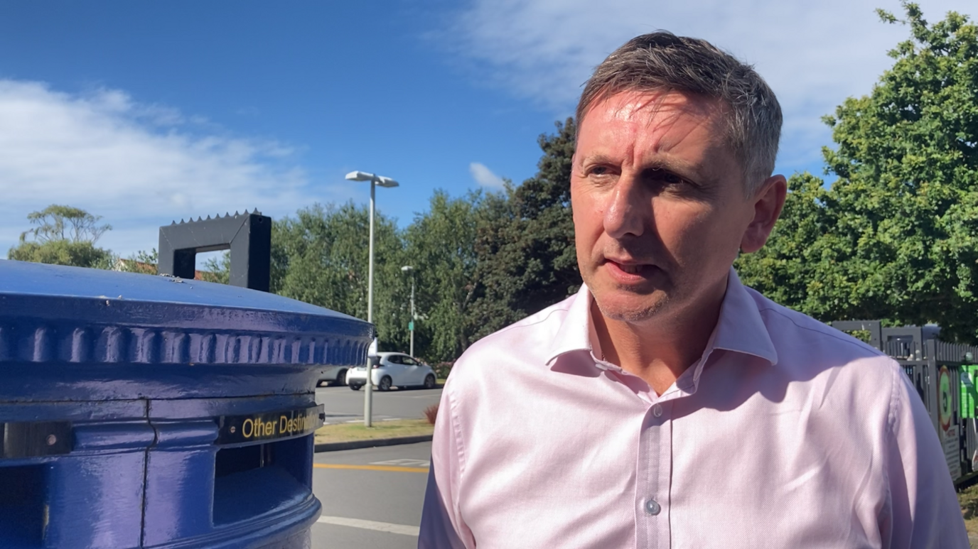 Steve Sheridan standing to the right of a blue post box. He is looking to the left and wearing a pink shirt. 