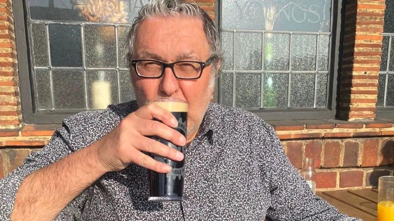 Kevin Tweedy sat on a bench outside a pub. He has grey hair, black glasses and is holding a pint of beer to his lips