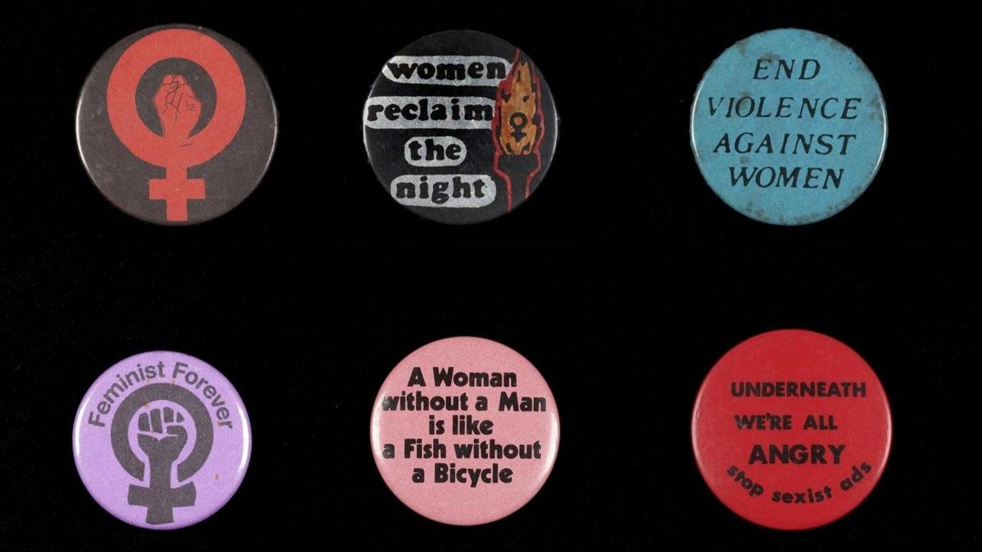 A selection of badges on display in the exhibition. They have slogans including "End violence against women" and "Feminist Forever".