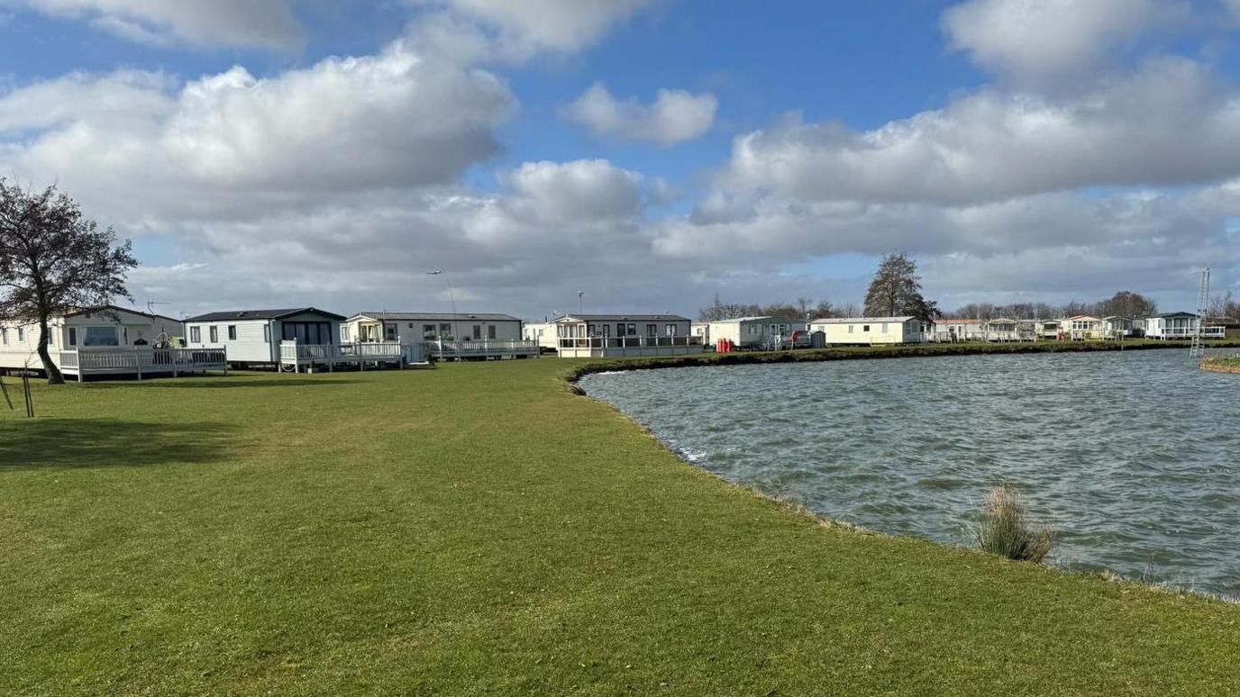 A row of white caravans next to a blue lake surrounded by grass, and two trees. The sky is blue with white fluffy clouds.