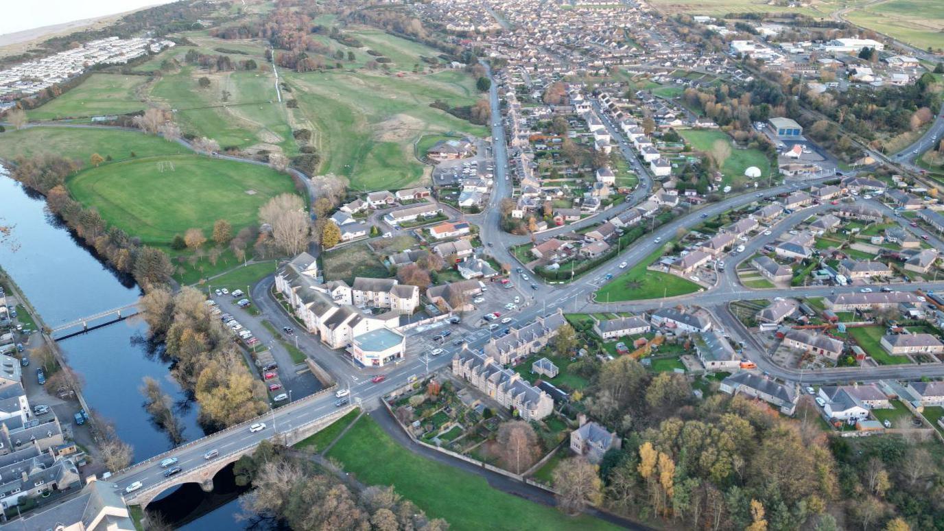 An aerial view of the A96 as it passes through Nairn. There is a bridge over a river and housing developments.