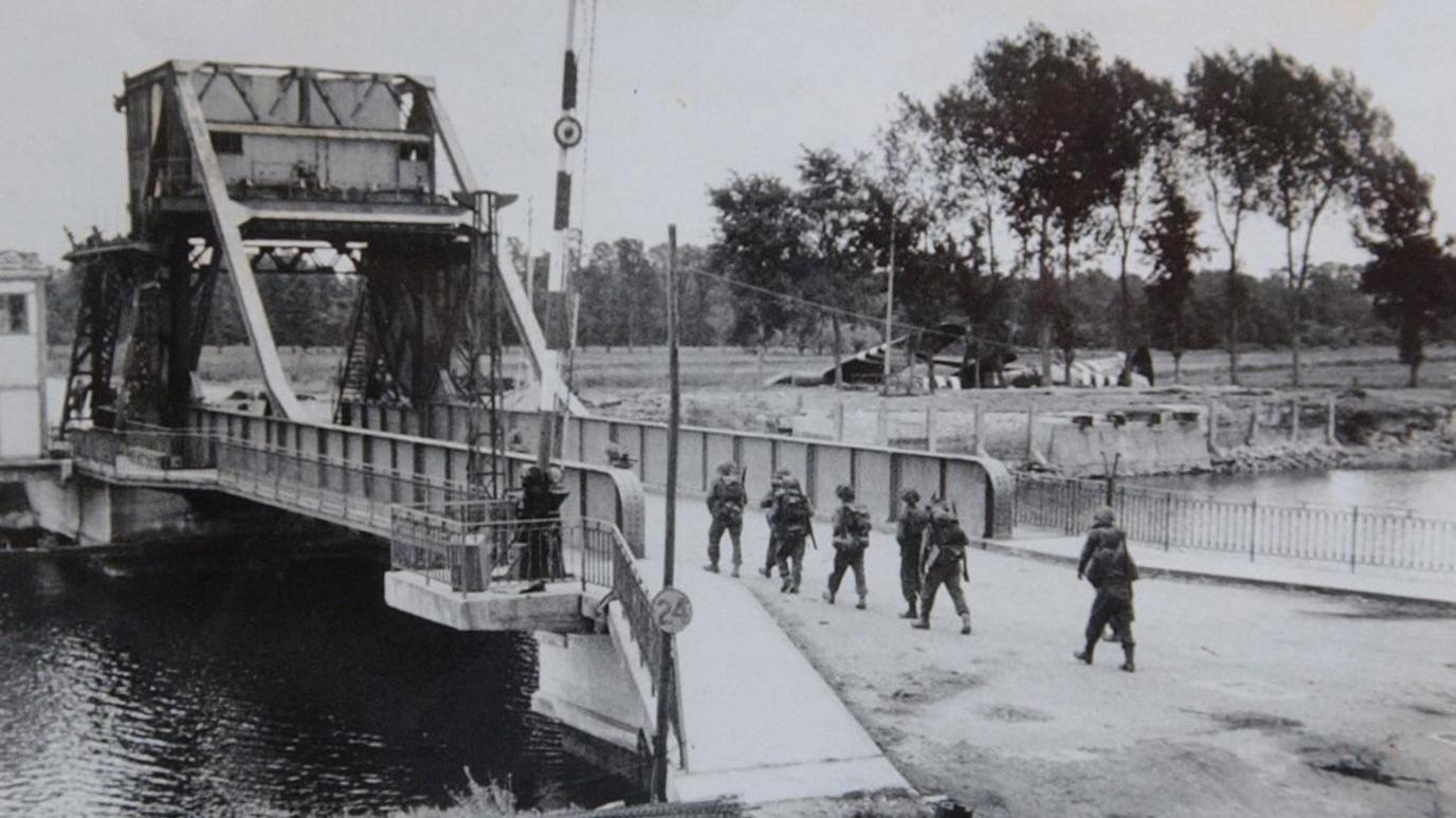 A black and white photo from 1944 of a metallic bridge over a river in France. Troops can be seen crossing the bridge. In the background gliders can be seen landed on the ground, slightly obscured by trees.