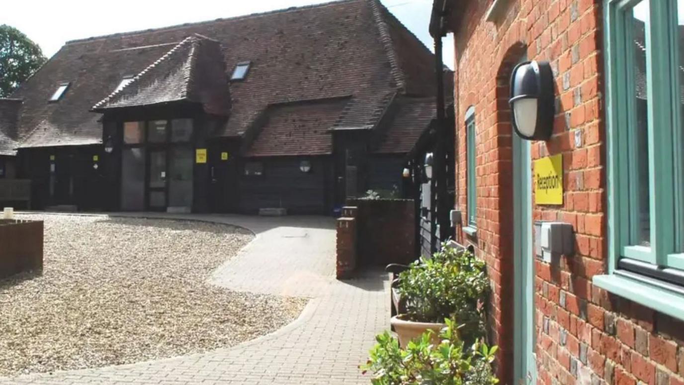 A dark farm-style building with tall and wide glass doors at the entrance sits at the end of a gravel path, with a red-brick building on the left-hand side which has a green windowsills and a green door and a yellow sign on the brick wall reads "reception"
