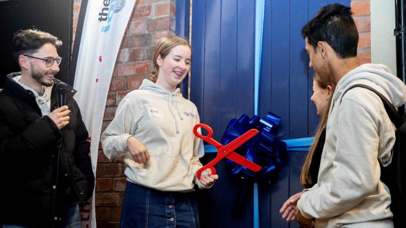 A woman in a grey hoodie prepares to cut a blue ribbon on a big blue door with some red scissors. Three people watch her.  