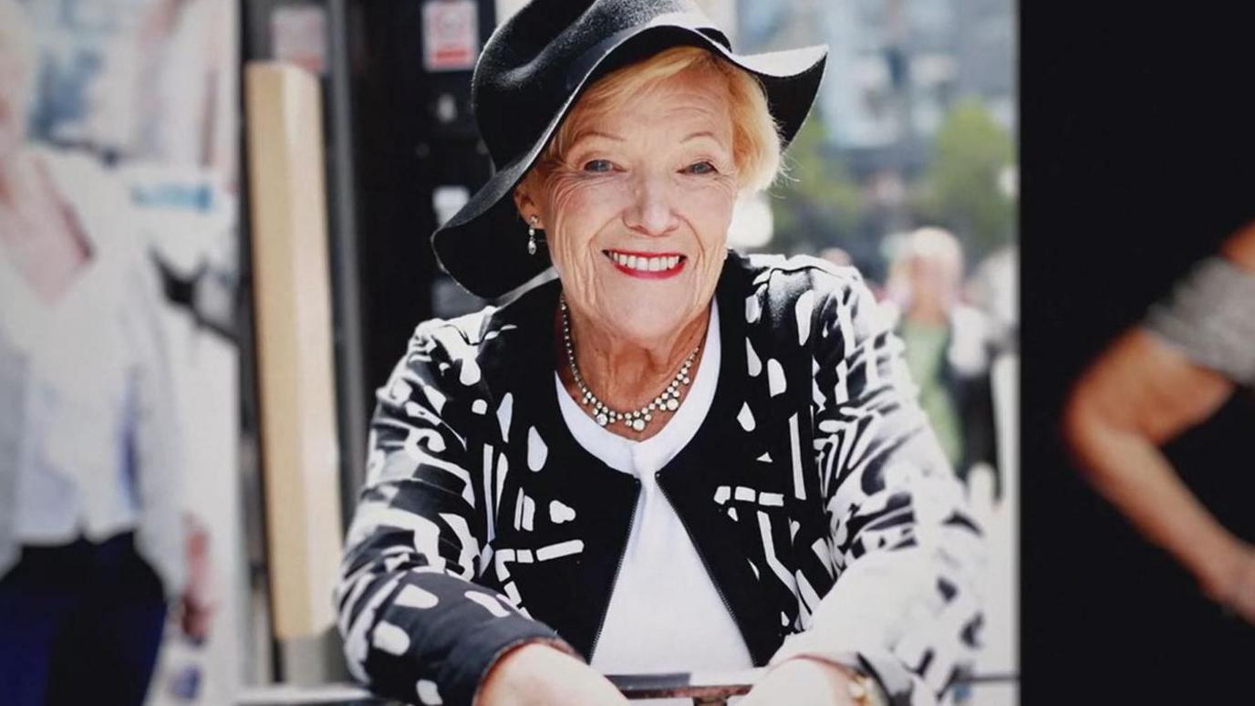 Old woman with blonde hair wearing a black hat and a black and white jacket 