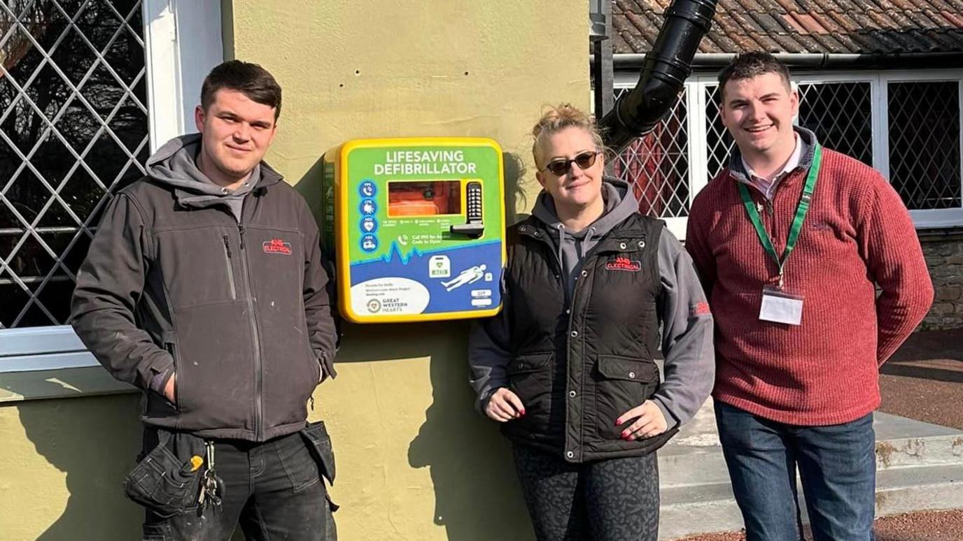 Two men and a woman standing next to a defibrillator which is attached to the outside wall of a pub.