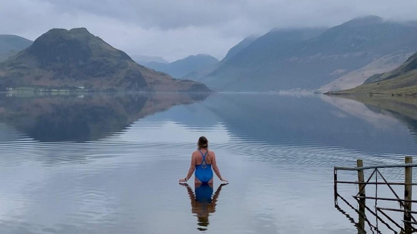 Sara Barnes stands in Crummock Water on a misty day. She wears a blue swimsuit and faces the mountains in the background.