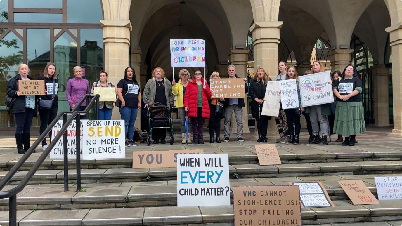 Parents and families of SEND children outside Guildhall in Northampton. They are holding placards and others are arranged on the steps in front of them. One reads "When will every child matter?"