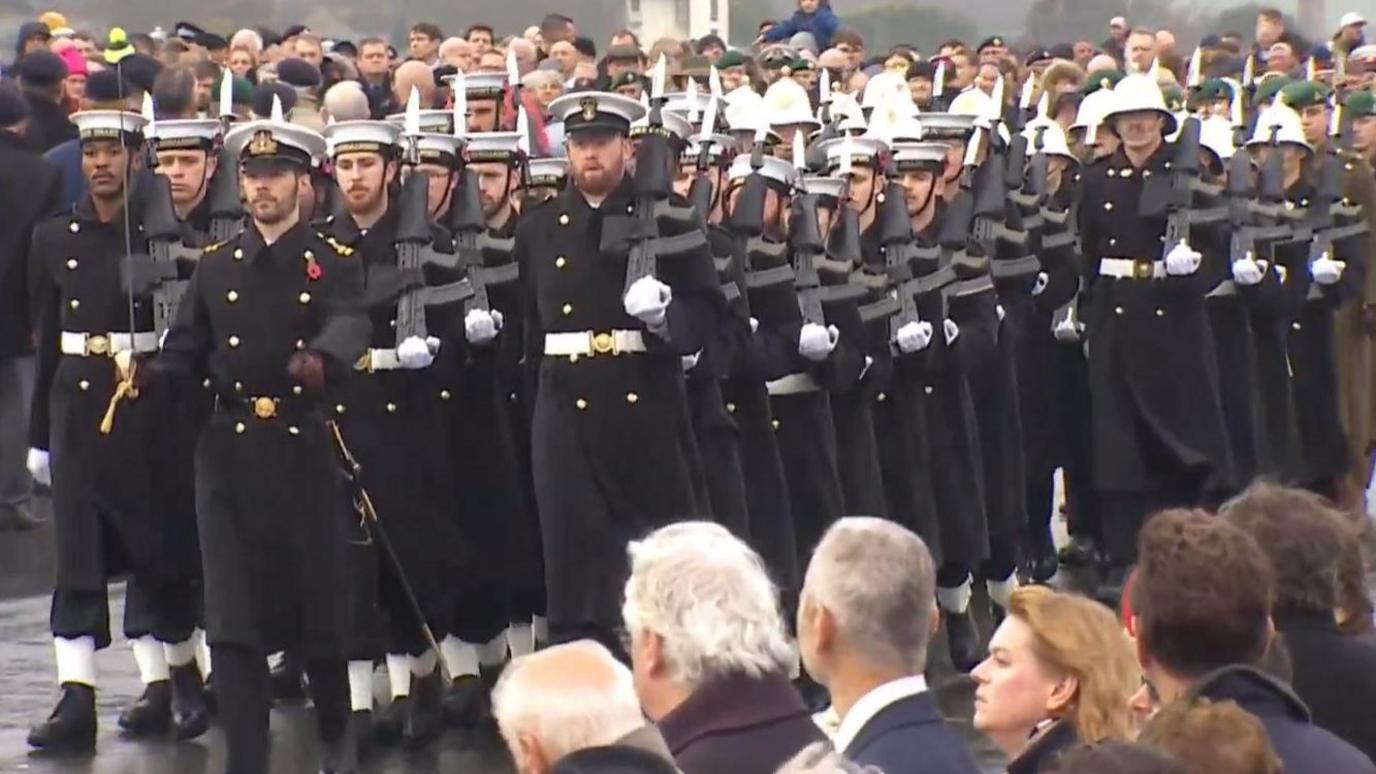 Members of the navy are seen marching down Plymouth Hoe. They are in black uniforms with white hats and holding a gun on their shoulders. They are marching between crowds.