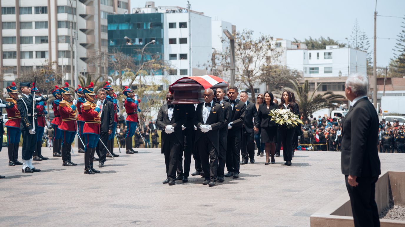 Several congressmen carry the coffin during the wake of former president Alberto Fujimori. His coffin is drapped with the national flag of Peru on the top of it and several members of his family walking pacing behind the coffin. Crowds and soldiers watch on from either side