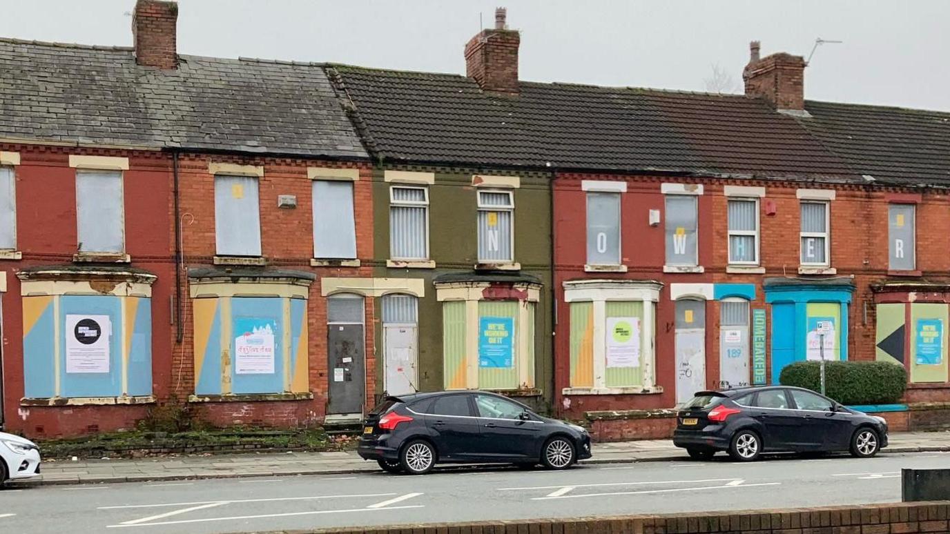 A wider view of the row of terraced houses with their windows and doors shuttered with metal sheets.