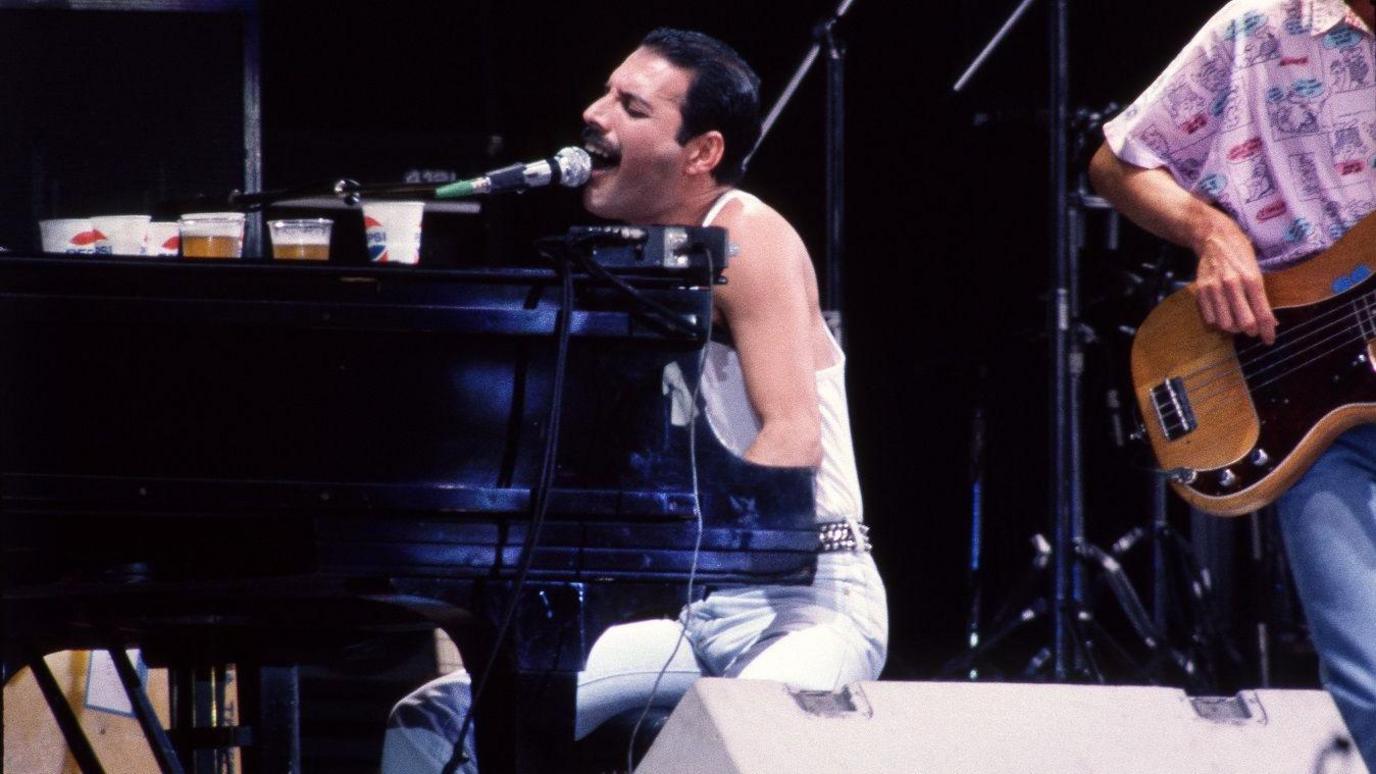 Freddie Mercury seated at a grand piano mid‑performance, leaning in to sing into the mic. There are several plastic drink cups lined up on the piano with a bassist visible in the background.