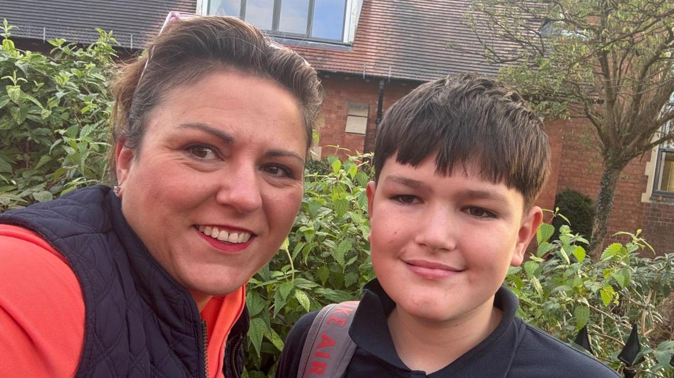 Jo, who has brown hair tied back into a ponytail and sunglasses on her head, takes a selfie alongside her son Jacob. He has dark brown hair too. They're stood in front of a leafy green hedge.