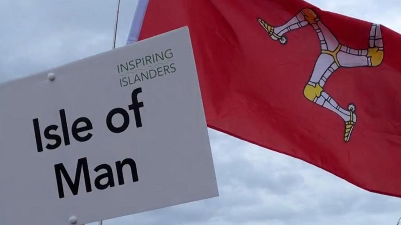 A white board on a brown stick is held in the sky, it reads Isle of Man. Behind it a red Manx flag also flies against the backdrop of a cloudy sky. It bears a yellow and white triskelion in its centre.