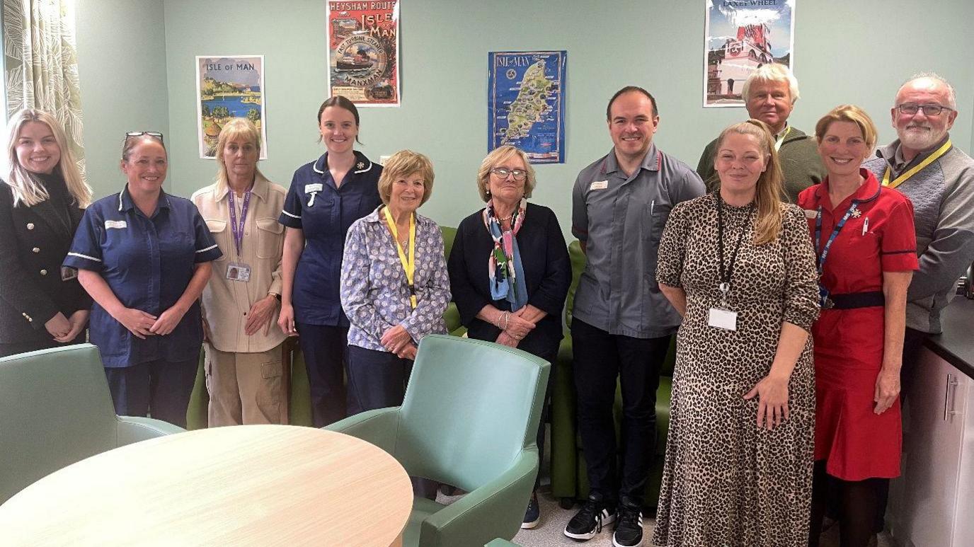 A group of 11 people smiling in a lounge, some are wearing nurses uniforms, and others have lanyards around their necks.