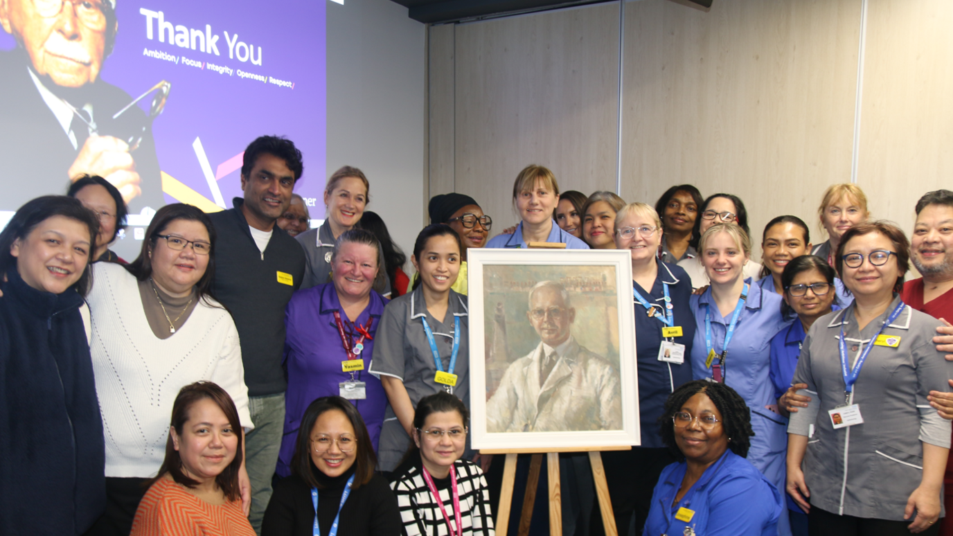 Staff from Guy’s and St Thomas’ NHS Foundation Trust gather round an easel displaying a portrait of Sir Harold Ridley. In the background a projected image on the wall features another image of him with the words 'thank you'