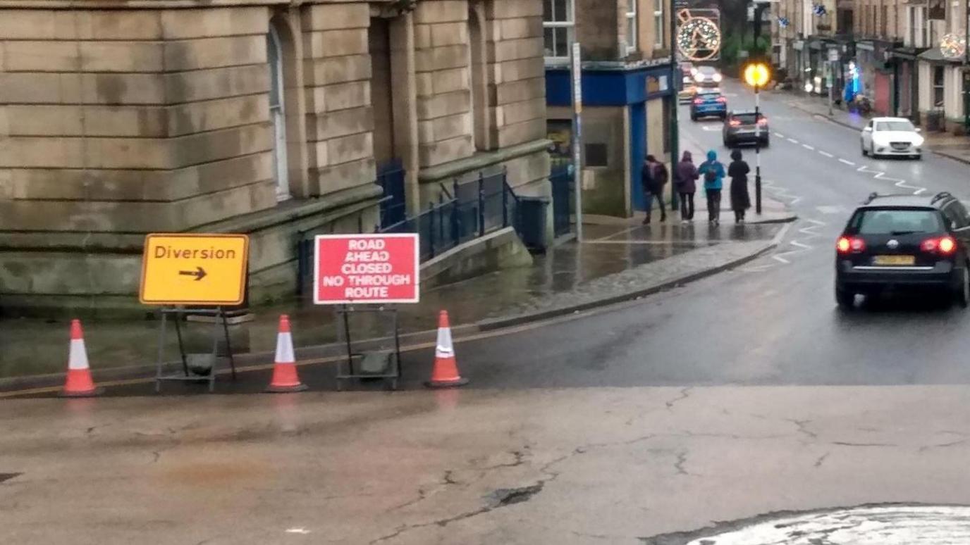 Diversion and road closure signs stand alongside cones on the corner of a roundabout. Cars travel in the background.