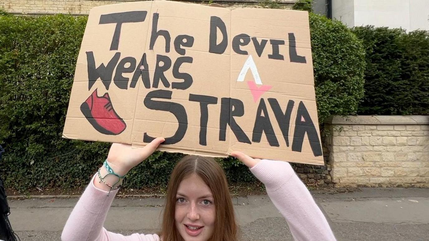 A girl holding up a sign saying 'The Devil Wears Strava'.