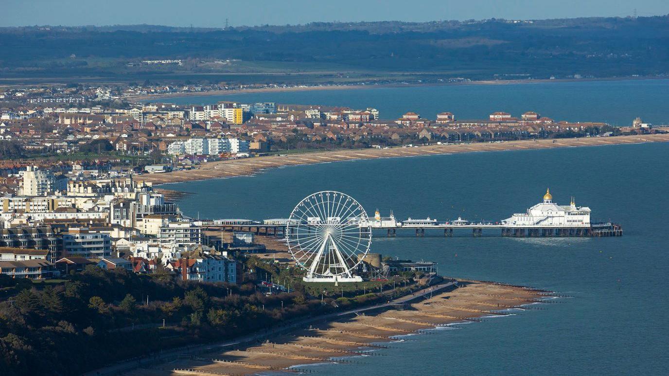 A view of the route to Eastbourne from Beachy Head, showing the coastline stretching around the grey-blue sea, with a white Ferris Wheel and a white pier and groups of buildings of different heights