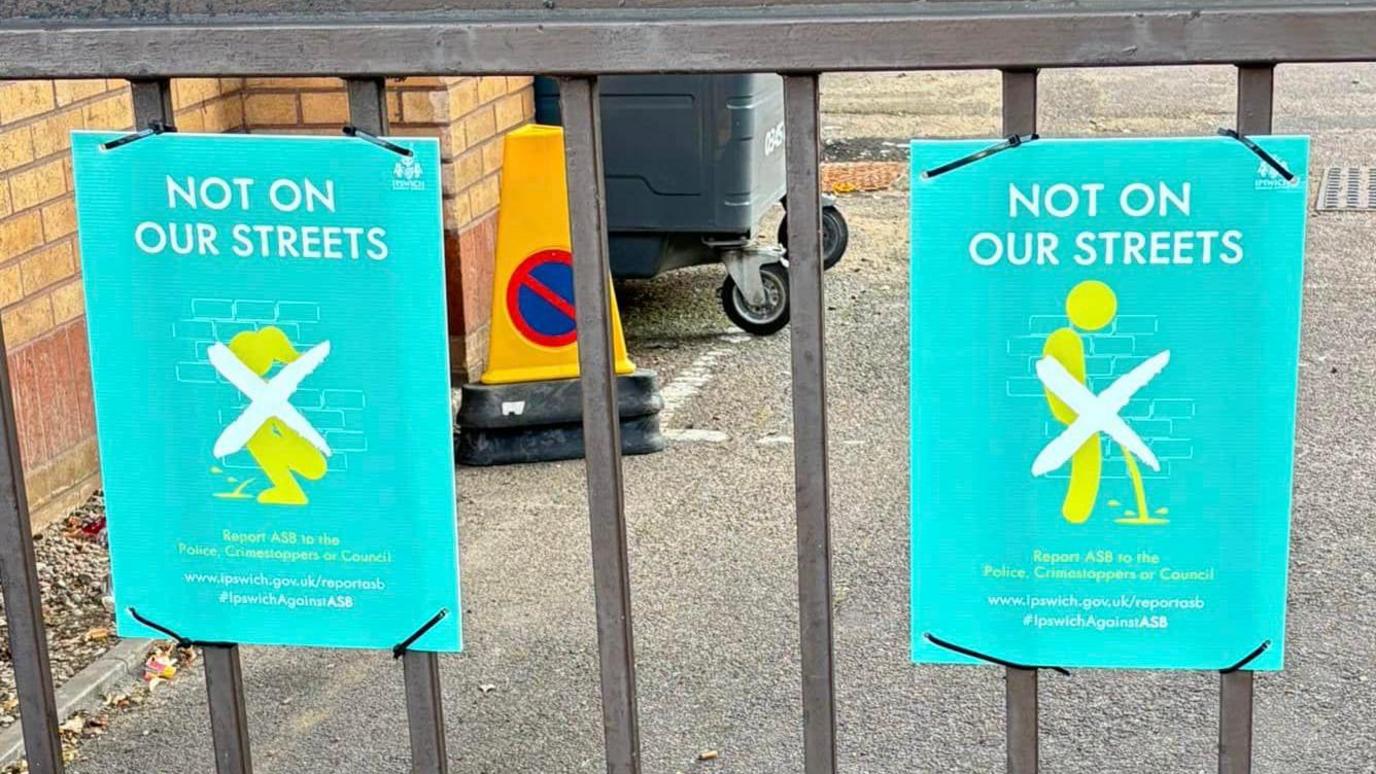 Two green posters attached to a gate, both with graphics showing people urinating and large X over the top and headline reading "Not On Our Streets"