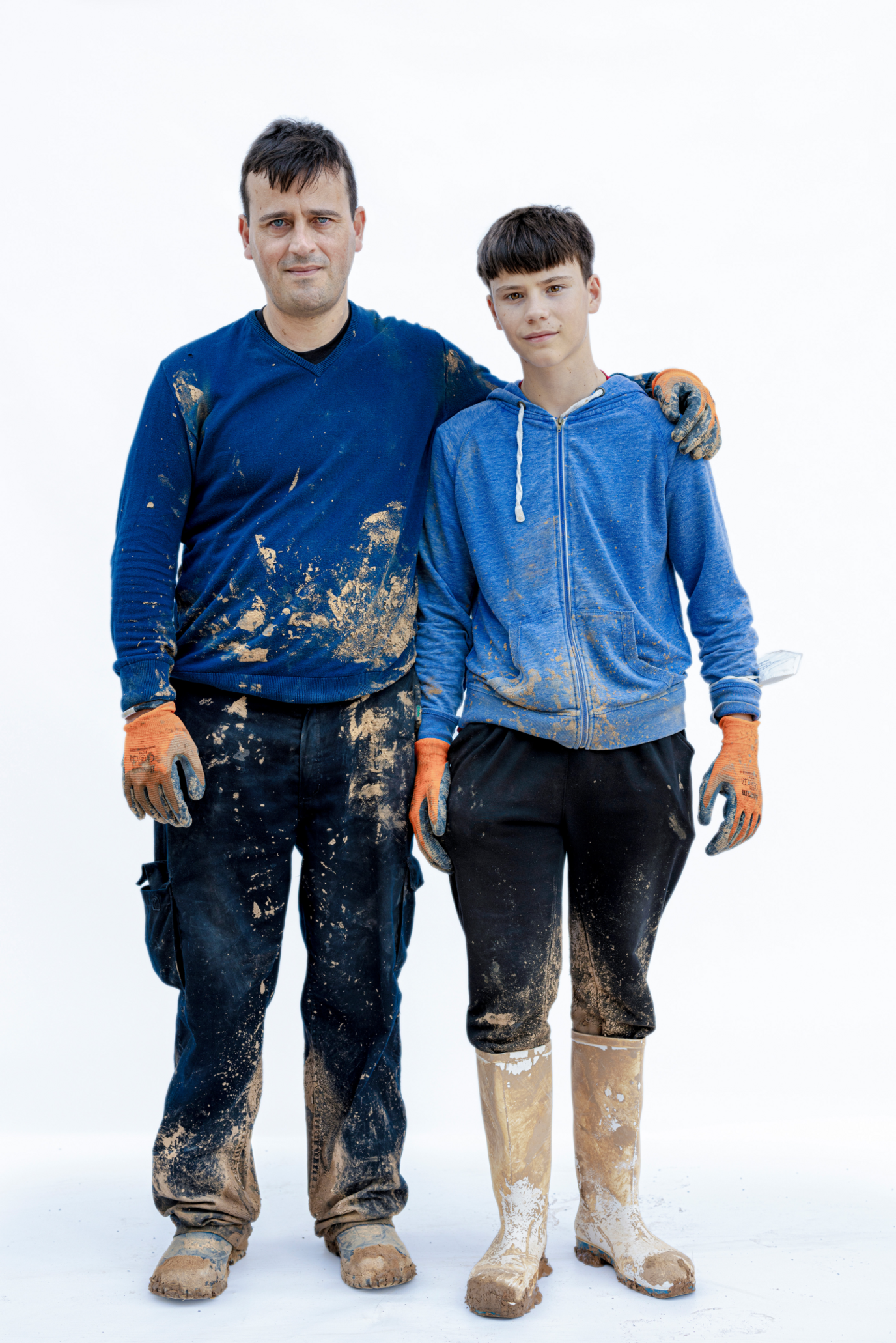 Image Description: A man has his arm around the shoulders of a teenage boy, both are staring at the viewer while standing in an empty space. both are wearing gloves and their clothes are covered in mud.