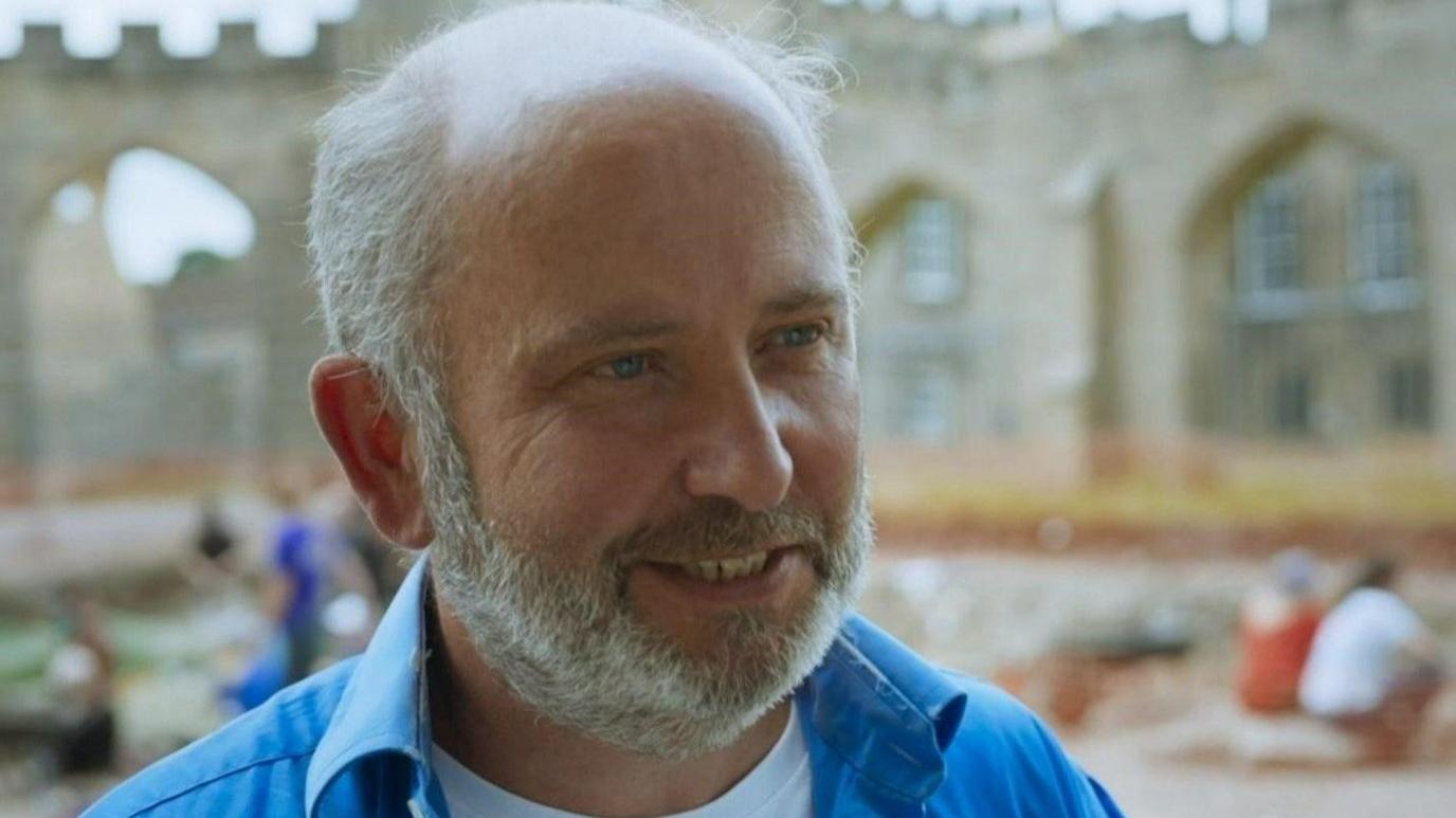 Professor Chris Gerrard is smiling during the excavation. He has straight white hair and beard. His eyes are blue and the top of his head in bald. He is wearing a blue unbuttoned shirt and a white t-shirt.  