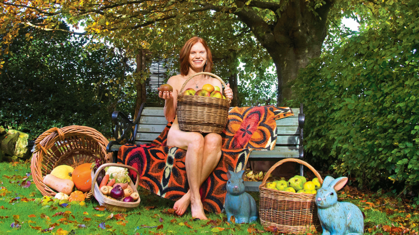 The image shows a naked woman sitting on a bench with baskets of fruit and vegetables around her. She has a basket of apples modestly placed on her lap. 