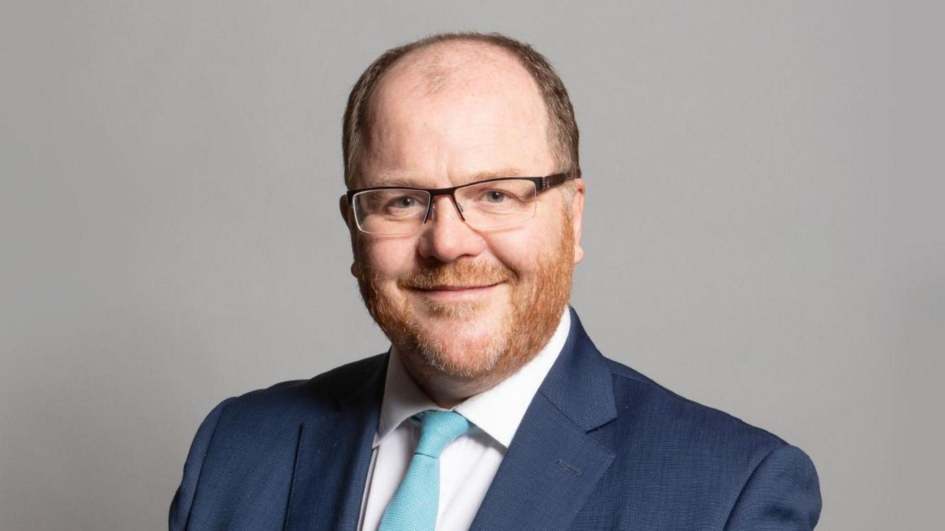 George Freeman is smiling at the camera. He's wearing glasses, a blue suit jacket, white shirt and light blue tie.