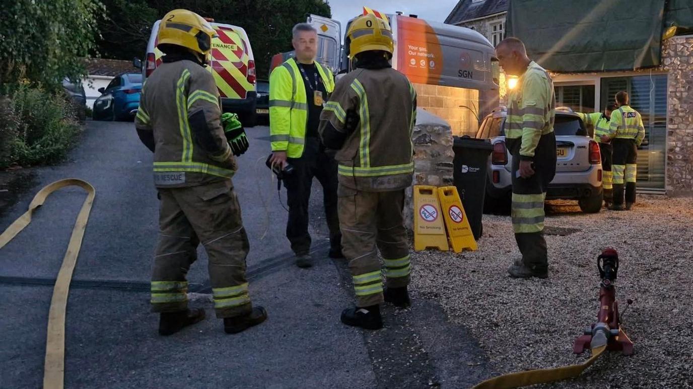 Fire crews and gas board engineers standing in a narrow lane at dusk. To the right of the picture is a house and two people wearing hi-viz peering at a silver car.