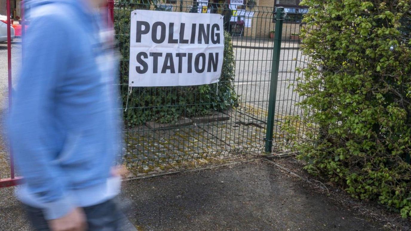 A polling station sign on a black gate as a man in a blue shirt walks past.