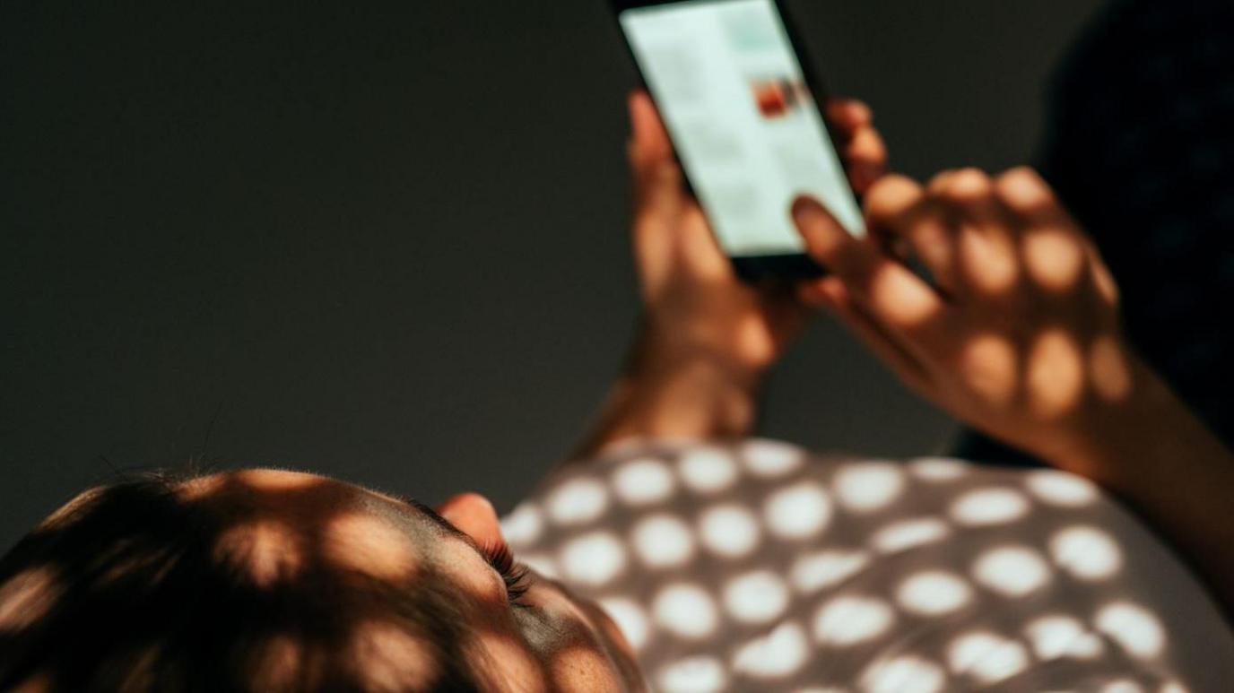 A woman is lying on a bed and holding a mobile phone. Light from a window is making a shining a pattern on her top. 