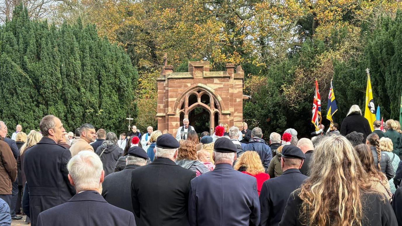 Rows of people in dark clothing, seen from behind with a number of flags and a small stone structure in front of them and trees in the distance
