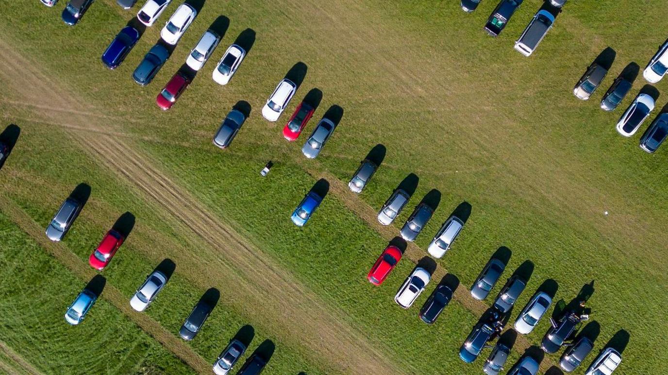 Aerial view of vehicles parked in a green field. The cars are red, blue, black and white.