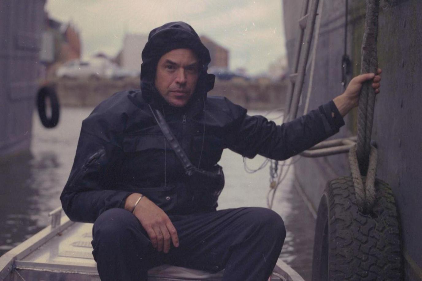 Artist Mark Pozlep, wearing a waterproof with the hood up, sits in a boat holding on to a rope.