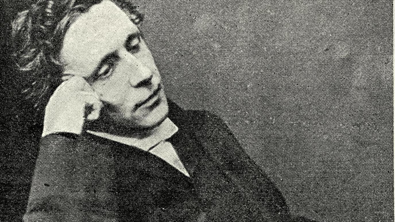 Archive photo of Lewis Carroll seated in a relaxed pose, wearing formal Victorian-era clothing including a dark suit and white shirt. He is resting one arm on the chair and the other near the face.