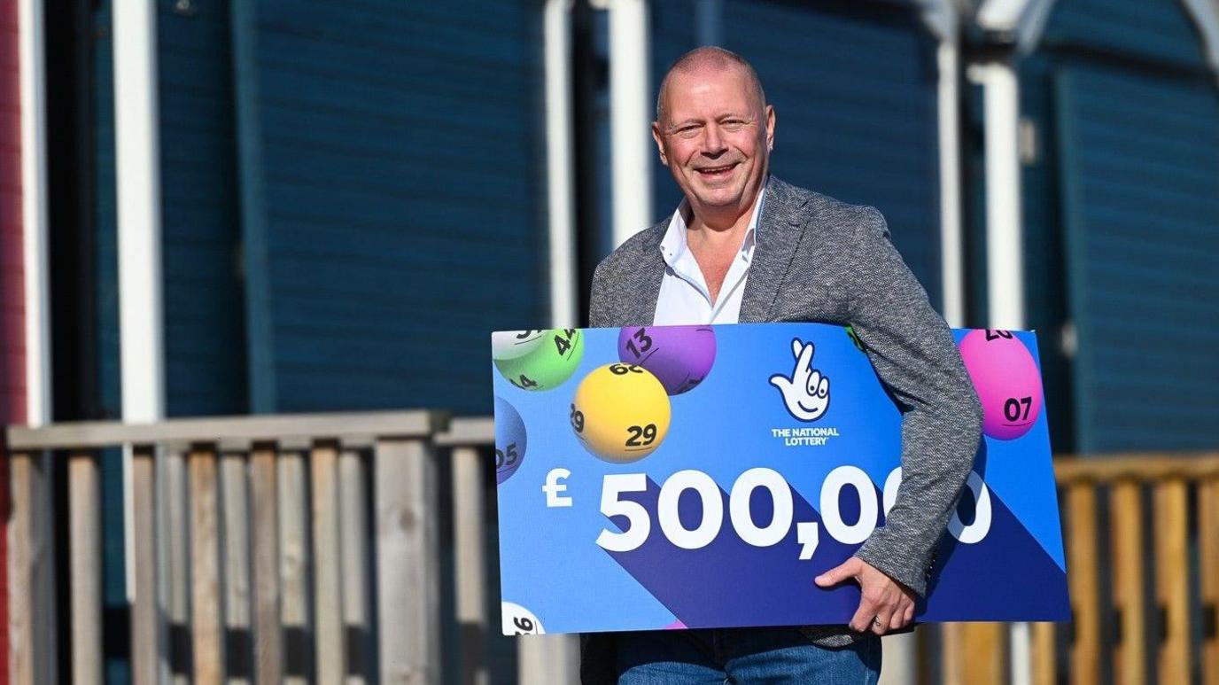 John Lingard is wearing a suit jacket and jeans, he is walking towards the camera and is smiling. He is carrying a blue placard which has National Lottery branding and says £500,000.