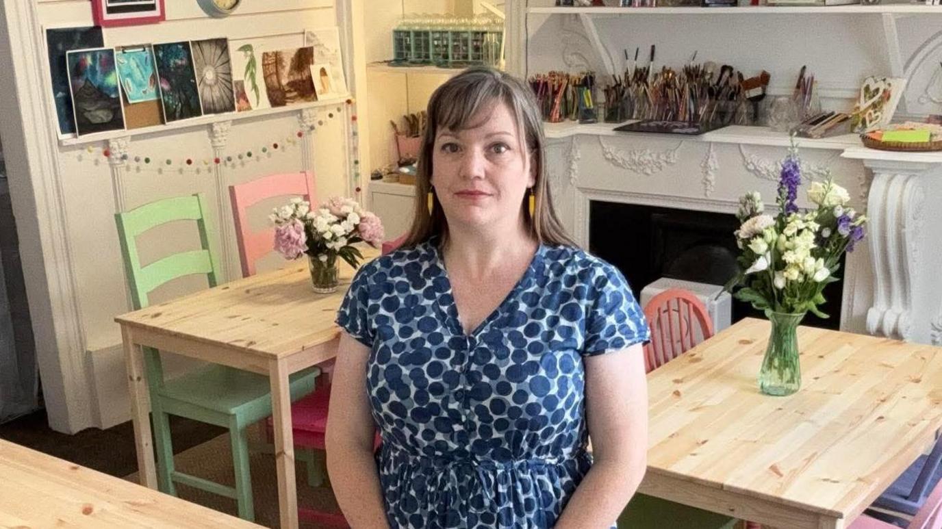 Carly is stood in the middle of a room with colourful art supplies in the background. She is wearing a blue spotted dress and yellow earrings. She has dark blonde hair and a fringe.