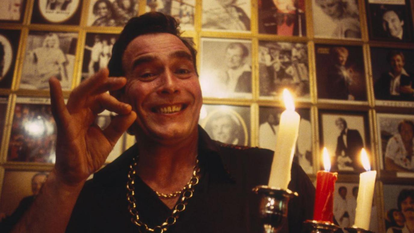 George pictured in 1993 holding an alight candelabra while wearing a black shirt and gold necklaces. He is smiling in front of a display of lots of black and white portraits. He has his fingers pushed together to make an "OK" gesture.